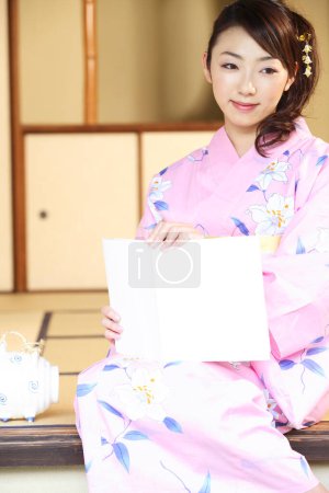 Téléchargez les photos : Une femme dans un kimono assis à la maison avec un livre - en image libre de droit