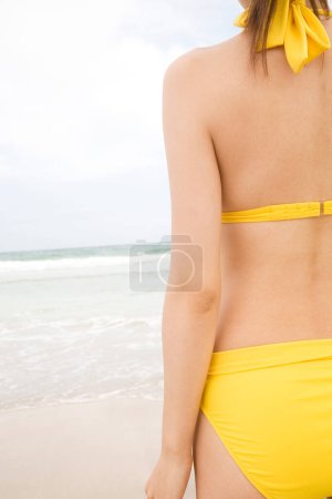 Foto de Foto recortada de la mujer con traje de baño amarillo de pie con su espalda en la playa - Imagen libre de derechos