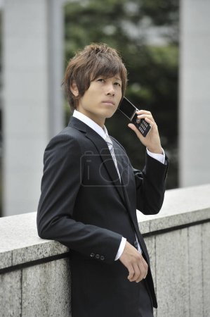Téléchargez les photos : Jeune asiatique homme d'affaires en costume parler sur téléphone mobile sur la rue - en image libre de droit