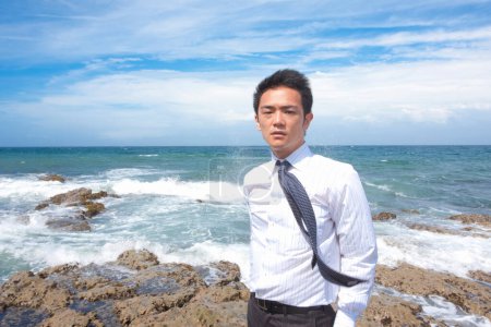 Téléchargez les photos : Portrait de beau adulte asiatique homme d'affaires sur le bord de la mer sur fond de ciel bleu - en image libre de droit