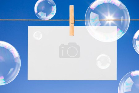 Téléchargez les photos : Feuille de papier vierge accrochée à la corde des vêtements et des bulles sur fond bleu ciel - en image libre de droit