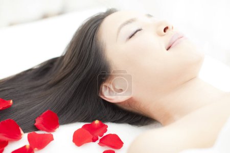 Téléchargez les photos : Belle femme asiatique avec les yeux fermés se détendre sur le lit avec des pétales de rose rouge dans le salon de spa. - en image libre de droit