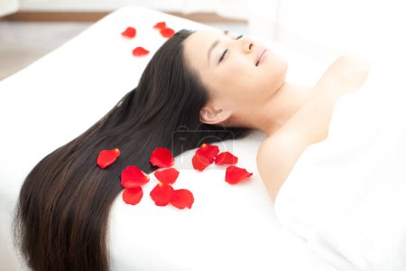 Foto de Hermosa mujer asiática con los ojos cerrados relajarse en la cama con pétalos de rosa roja en el salón de spa. - Imagen libre de derechos