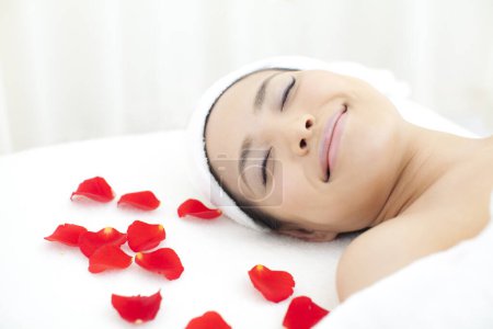 Téléchargez les photos : Belle femme asiatique avec les yeux fermés et serviette blanche sur la tête posée sur le lit avec des pétales de rose rouge dans le salon de spa. - en image libre de droit