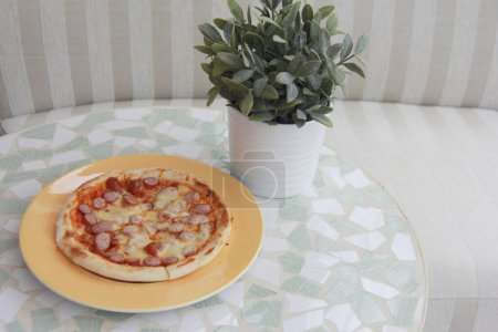 Photo for Delicious pizza with sausages and cheese near green plant on table - Royalty Free Image