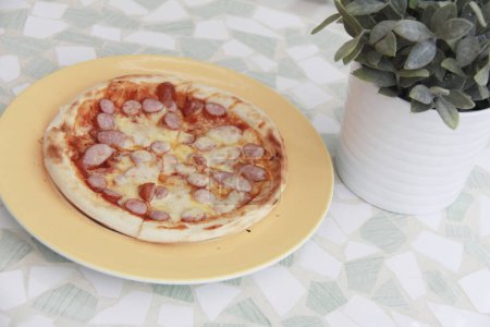 Photo for Delicious pizza with sausages and cheese near green plant on table - Royalty Free Image
