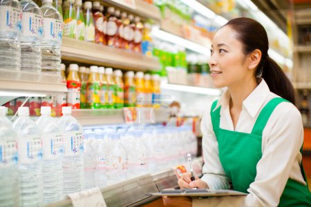 Photo for Cheerful Japanese woman in green apron. Smiling worker with clipboard in grocery store - Royalty Free Image