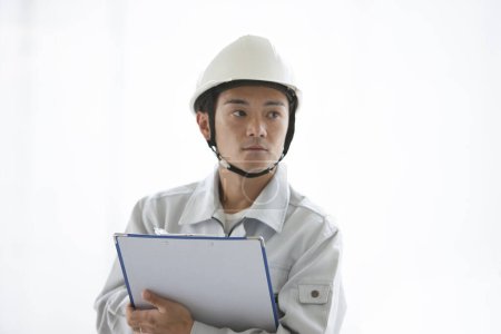Photo for Portrait of handsome young Japanese builder in uniform inspecting construction site with clipboard - Royalty Free Image