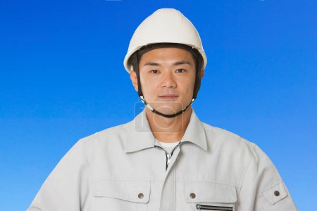 Téléchargez les photos : Travailleur japonais confiant dans la construction. jeune homme avec casque sur fond bleu - en image libre de droit