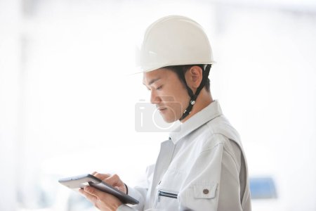 Foto de Retrato de joven constructor japonés guapo en uniforme en el sitio de construcción utilizando tableta digital - Imagen libre de derechos