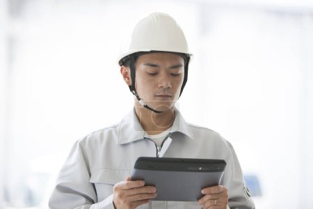 Téléchargez les photos : Portrait du beau jeune constructeur japonais en uniforme sur le chantier à l'aide d'une tablette numérique - en image libre de droit
