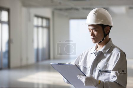 Téléchargez les photos : Portrait de beau jeune constructeur japonais en uniforme inspectant chantier de construction avec presse-papiers - en image libre de droit
