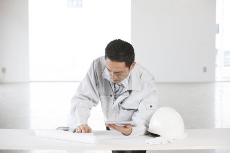 Téléchargez les photos : Portrait du beau jeune constructeur japonais en uniforme sur le chantier à l'aide d'une tablette numérique - en image libre de droit