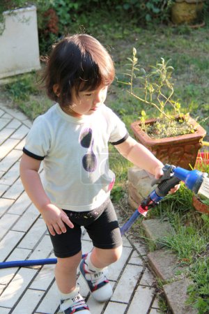 Téléchargez les photos : Mignon tout-petit asiatique fille avec un tuyau arrosage plantes dans jardin - en image libre de droit