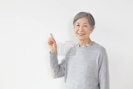 Foto de Mujer mayor sonriendo a la cámara y apuntando hacia arriba con el dedo sobre fondo blanco - Imagen libre de derechos