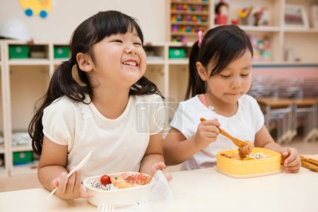 Photo for Two asian schoolgirls eating healthy food from their lunchboxes in classroom - Royalty Free Image