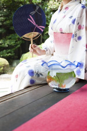 Photo for Young Japanese woman with paper fan and aquarium sitting on engawa and looking at garden - Royalty Free Image