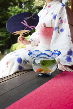 Téléchargez les photos : Jeune femme japonaise avec ventilateur de papier et aquarium assis sur engawa et regardant le jardin - en image libre de droit