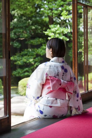 Téléchargez les photos : Vue arrière de la femme japonaise portant kimono assis sur engawa et regardant le jardin - en image libre de droit