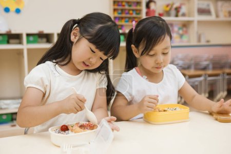 two asian schoolgirls eating healthy food from their lunchboxes in classroom