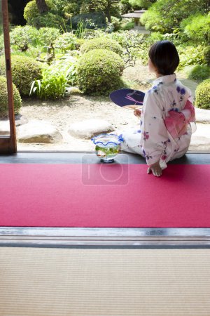 Photo for Young Japanese woman with paper fan and aquarium sitting on engawa and looking at garden - Royalty Free Image