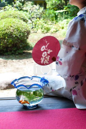 Téléchargez les photos : Jeune femme japonaise avec ventilateur de papier et aquarium assis sur engawa et regardant le jardin - en image libre de droit