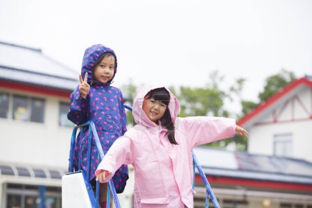 zwei süße asiatische Mädchen in Regenmänteln, die an einem bewölkten Tag auf dem Spielplatz spielen 