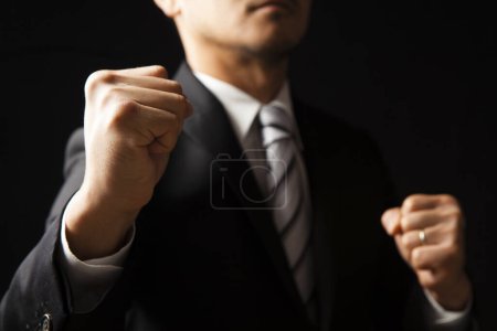 Téléchargez les photos : Asiatique homme d'affaires en costume noir et chemise blanche montrant poings sur fond noir - en image libre de droit