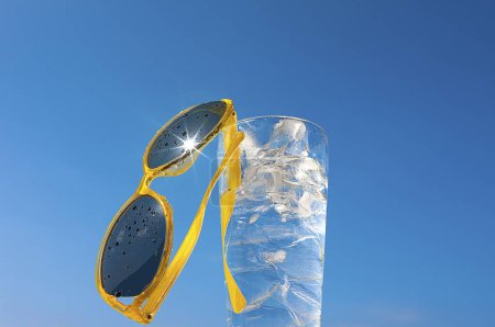 Foto de Vaso de agua con cubitos de hielo y gafas de sol amarillas en el fondo del cielo azul, concepto de vacaciones de verano y relajación en un día soleado, espacio para copiar - Imagen libre de derechos