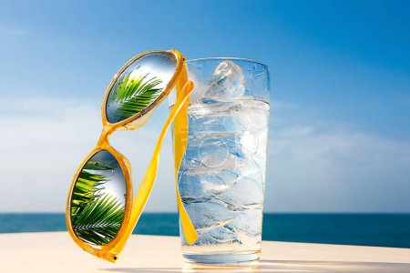 Foto de Vaso de agua con cubitos de hielo y gafas de sol amarillas en el fondo del mar, concepto de vacaciones de verano y relajación en un día soleado, espacio para copiar - Imagen libre de derechos