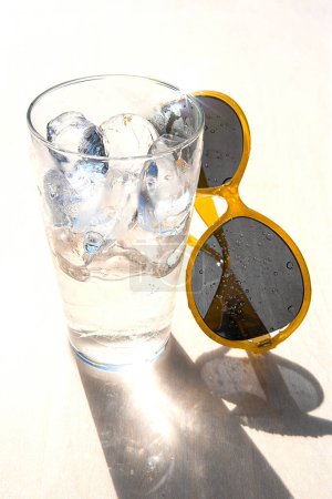 Photo for Glass of water with ice cubes and yellow sunglasses on white background, concept of summer vacation and relaxation in a sunny day, copy space - Royalty Free Image