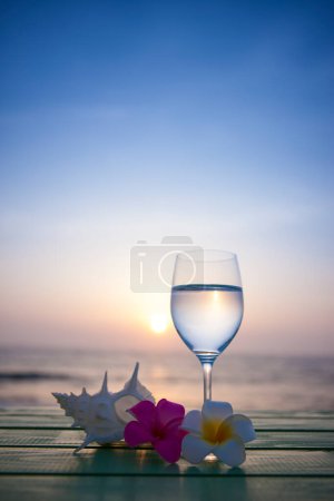 Téléchargez les photos : Fond d'été avec des fleurs de plumeria, coquille et verre à vin avec de l'eau sur la plage - en image libre de droit