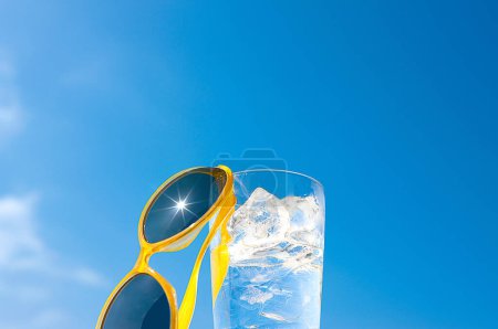 Foto de Vaso de agua con cubitos de hielo y gafas de sol amarillas en el fondo del cielo azul, concepto de vacaciones de verano y relajación en un día soleado, espacio para copiar - Imagen libre de derechos