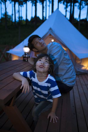 Foto de Asiático chico y su padre mirando arriba sentado en la mesa cerca de tienda de campaña mientras acampa en el bosque - Imagen libre de derechos