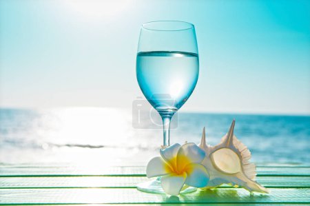 Téléchargez les photos : Fond d'été avec fleur de plumeria, coquille et verre à vin avec de l'eau sur la plage - en image libre de droit