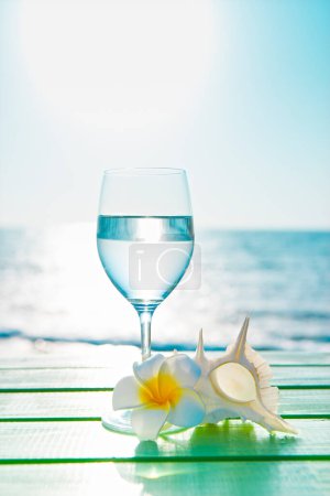 Téléchargez les photos : Fond d'été avec fleur de plumeria, coquille et verre à vin avec de l'eau sur la plage - en image libre de droit