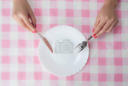 Foto de Vista superior de las manos femeninas sosteniendo tenedor y cuchillo sobre blanco plato vacío en la mesa con mantel a cuadros - Imagen libre de derechos