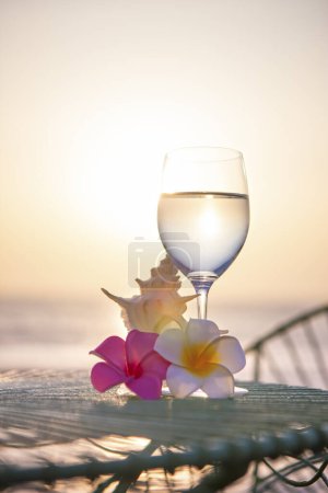 Téléchargez les photos : Fond d'été avec des fleurs plumeria, coquille et verre à vin avec de l'eau sur la plage à la lumière du coucher du soleil - en image libre de droit
