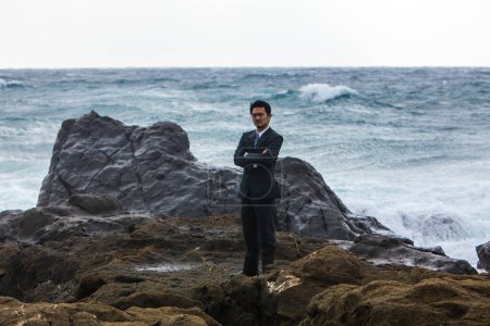 Foto de Joven asiático hombre de negocios en negro traje caminando en rocoso playa - Imagen libre de derechos