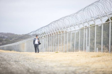 Téléchargez les photos : Asiatique homme d'affaires en costume formel promenades près de clôture - en image libre de droit