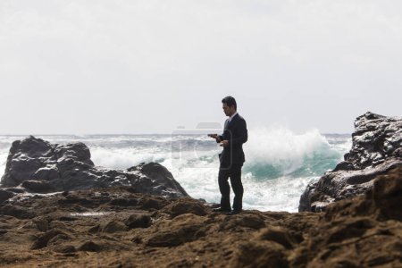 Téléchargez les photos : Jeune homme d'affaires asiatique en costume noir marchant sur une plage rocheuse et utilisant un smartphone - en image libre de droit