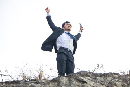 Foto de Joven hombre de negocios asiático levantando brazos con teléfono inteligente en la playa rocosa - Imagen libre de derechos