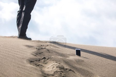 Téléchargez les photos : Section basse de l'homme d'affaires marchant à travers le téléphone qui est coincé dans le sable - en image libre de droit
