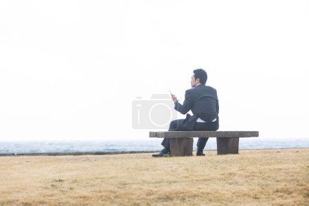 Téléchargez les photos : Asiatique homme d'affaires en costume formel assis sur banc et en utilisant téléphone, vue arrière - en image libre de droit