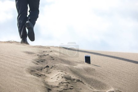 Photo for Low section of businessman walking through phone which is stuck in sand - Royalty Free Image