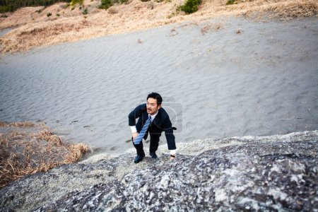 Foto de Asiático hombre de negocios escalada rocas en arenosa playa - Imagen libre de derechos