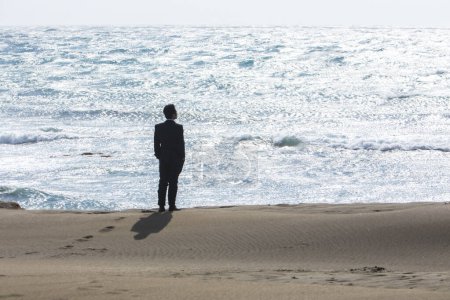 Téléchargez les photos : Jeune homme d'affaires asiatique en costume noir marchant sur la plage - en image libre de droit