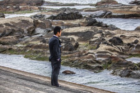 Photo for Young asian businessman in black suit walking on rocky beach - Royalty Free Image