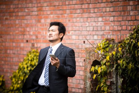 Foto de Asiático hombre de negocios usando móvil cerca de ladrillo pared en ciudad - Imagen libre de derechos