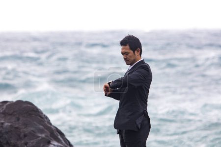 Téléchargez les photos : Jeune homme d'affaires asiatique en costume noir vérifier le temps tout en marchant sur la plage rocheuse - en image libre de droit